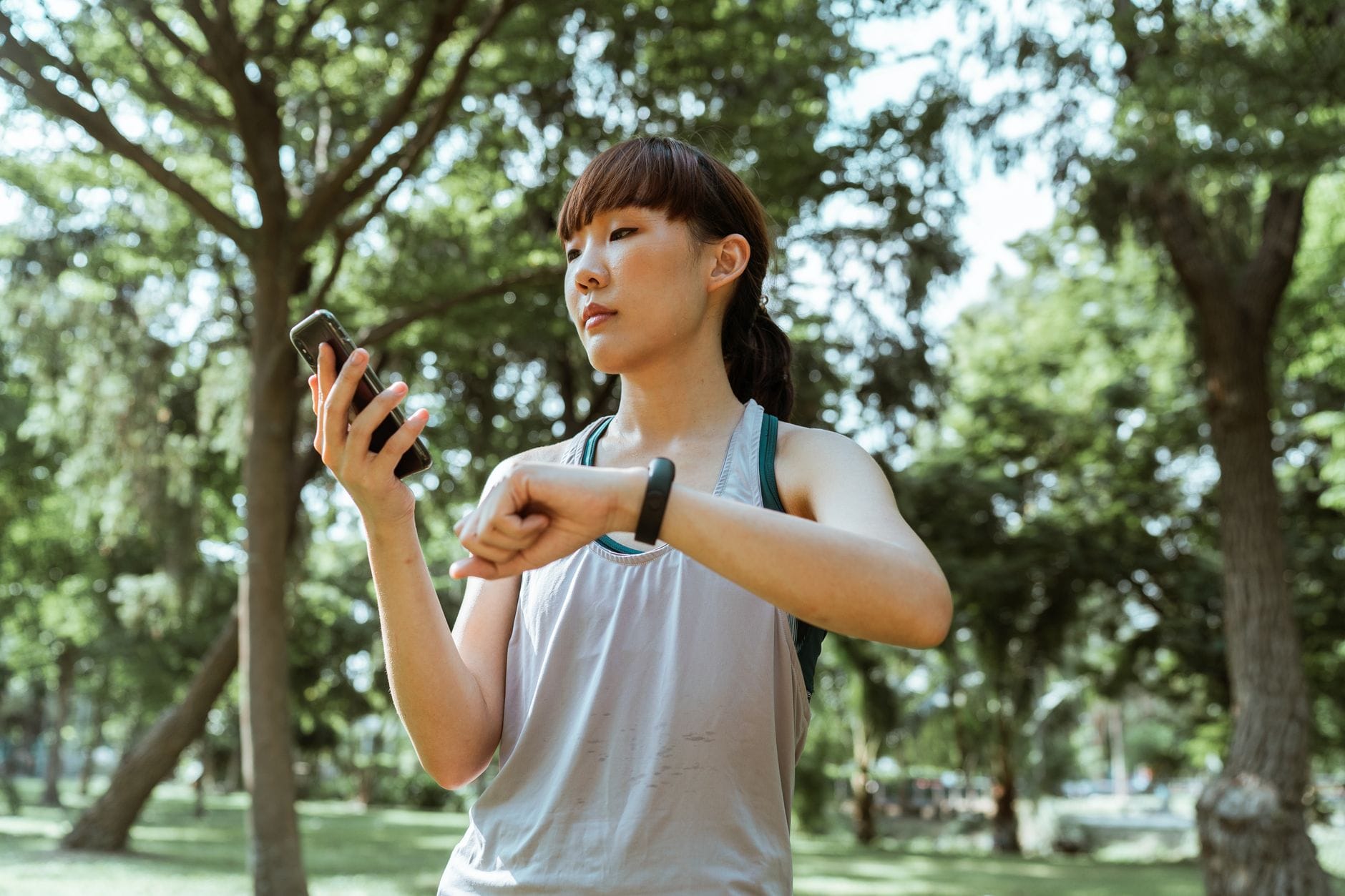 Młoda Azjatka w stroju sportowym sprawdza smartfon i opaskę fitness w parku